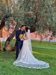 posi si baciano sotto un albero di ulivo, la sposa tiene in mano un bouquet di fiori.