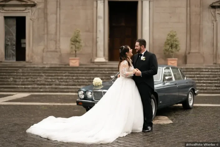 Coppia di sposi in abiti eleganti si abbraccia davanti alla chiesa, con un’auto d’epoca alle spalle, matrimonio a Viterbo