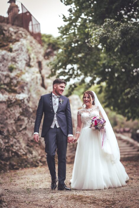 Coppia di sposi in abiti principeschi si tiene per mano camminando in un paesaggio naturale il giorno del loro matrimonio