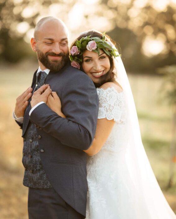 Sposa sorride abbracciando lo sposo al tramonto, indossando un abito in pizzo e corona di fiori, matrimonio a Viterbo