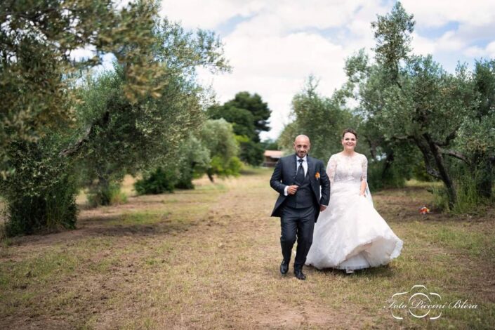 Coppia di sposi che corre mano nella mano in un uliveto, sorridendo felici.