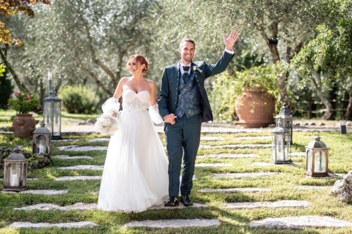 Coppia di sposi che salutano sorridendo, mano nella mano, circondati dagli invitati."